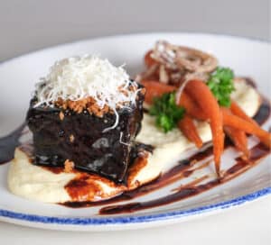 Photo of 48 hour short rib on plate with creamy polenta, natural jus, roasted carrot, crispy shallot, parmesan gremolata.