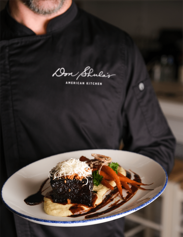 Photo of chef holding plate with 48 hour short rib, creamy polenta, roasted carrots, crispy shallot, and parmesan gremolata.