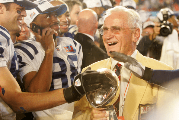 Vintage photo of smiling Don Shula in yellow sports coat holding Superbowl trophy.